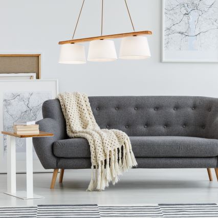 Trojramenné závesné svietidlo s drevenou konštrukciou a bielymi tienidlami visí nad sivou pohovkou s háčkovanou dekou. Pohovka stojí v svetlej miestnosti s obrázkami na stenách.