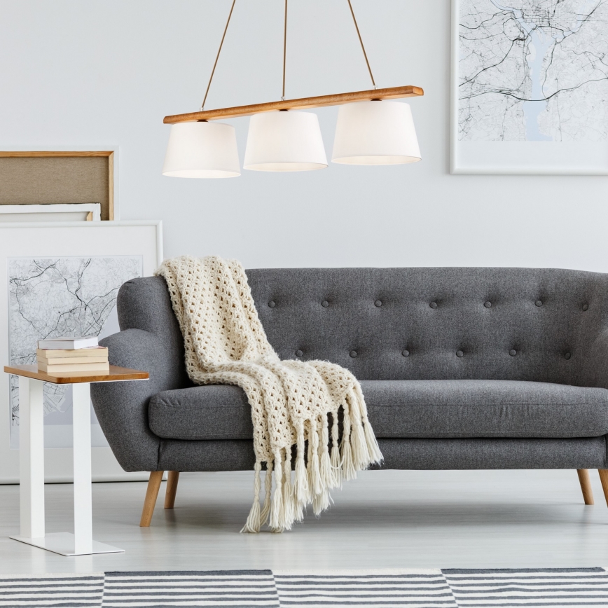 Trojramenné závesné svietidlo s drevenou konštrukciou a bielymi tienidlami visí nad sivou pohovkou s háčkovanou dekou. Pohovka stojí v svetlej miestnosti s obrázkami na stenách.