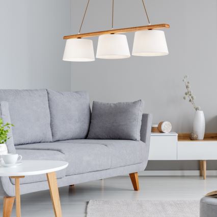 Trojramenné drevené závesné svietidlo s textilnými tienidlami visí nad sivou pohovkou v svetlej miestnosti. Pohovka je doplnená vankúšmi. V pozadí je vidieť biely nábytok a dekorácie.