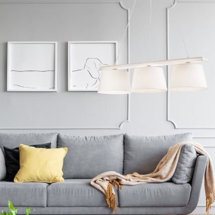 Trojramenné závesné svietidlo s bielymi tienidlami visí nad sivou pohovkou. Pohovka je doplnená vankúšmi a svetlohnedou dekou. Na stene za pohovkou sú dva obrazy.