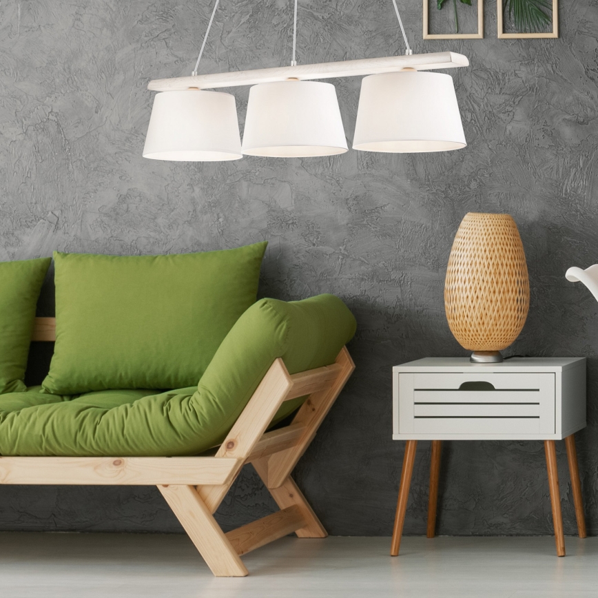 Trojramenné závesné svietidlo s bielymi tienidlami a drevenou konštrukciou visí nad zelenou rozkladacou sedačkou v interiéri. Vedľa sedačky stojí malý biely nočný stolík s pletenou vázičkou.