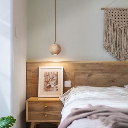 Drevená guľatá lampa visí nad drevenou posteľou s nočným stolíkom. Na stolíku je obraz mačky. Stena je z dreva a je na nej zavesená makramé.