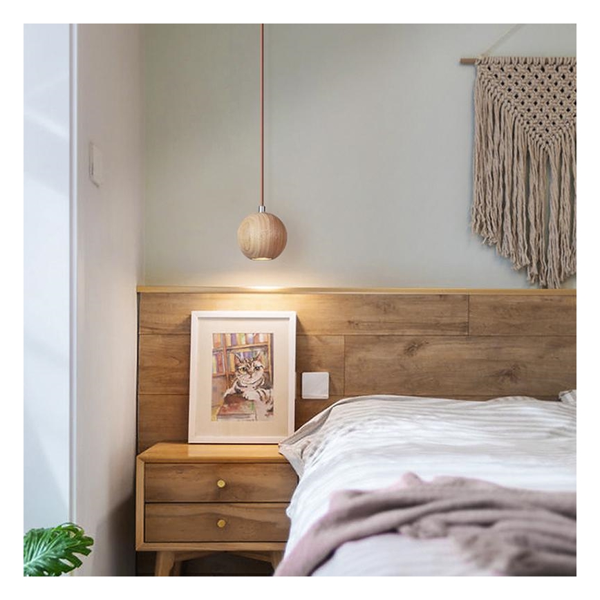 Drevená guľatá lampa visí nad drevenou posteľou s nočným stolíkom. Na stolíku je obraz mačky. Stena je z dreva a je na nej zavesená makramé.