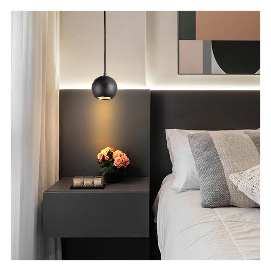 Čierna guľová lampa svieti v spálni. Lampa je zavesená nad tmavosivým nočným stolíkom s kvetmi a sviečkami. Vedľa je posteľ s bielou posteľnou bielizňou a vankúšmi.