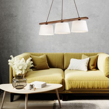 Trojramenné závesné svietidlo s drevenou konštrukciou a bielymi tienidlami visí nad žltou pohovkou v obývacej izbe. Na pohovke sú vankúše. Na konferenčnom stolíku pred pohovkou sa nachádzajú knihy a váza s kvetmi.