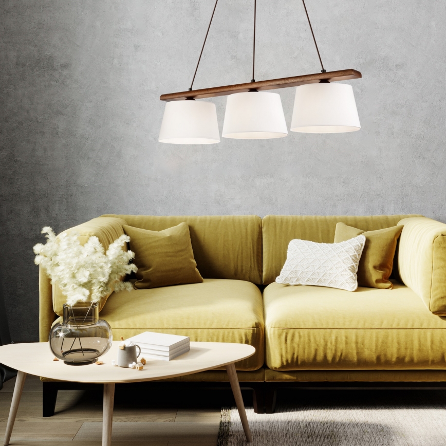 Trojramenné závesné svietidlo s drevenou konštrukciou a bielymi tienidlami visí nad žltou pohovkou v obývacej izbe. Na pohovke sú vankúše. Na konferenčnom stolíku pred pohovkou sa nachádzajú knihy a váza s kvetmi.
