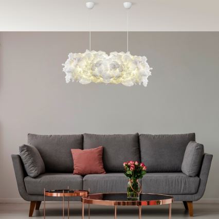 Biela oblaková lampa svieti nad sivou sedačkou. Sedačka je doplnená ružovými vankúšmi a vázou s kvetmi. V miestnosti sú aj dva odkladacie stolíky.