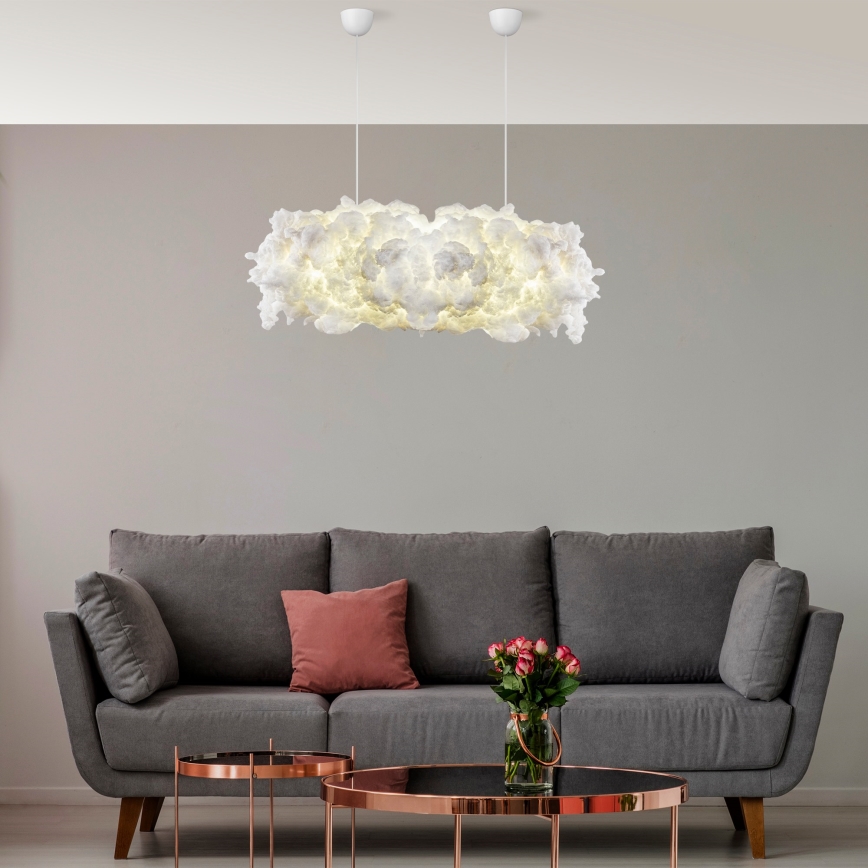 Biela oblaková lampa svieti nad sivou sedačkou. Sedačka je doplnená ružovými vankúšmi a vázou s kvetmi. V miestnosti sú aj dva odkladacie stolíky.