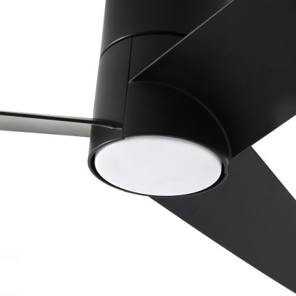 Čierny stropný ventilátor s tromi lopatkami sa nachádza pred bielym difúzorom svetla. Lopatky sú dlhé a úzke, pripevnené k valcovitému čiernemu telu ventilátora.