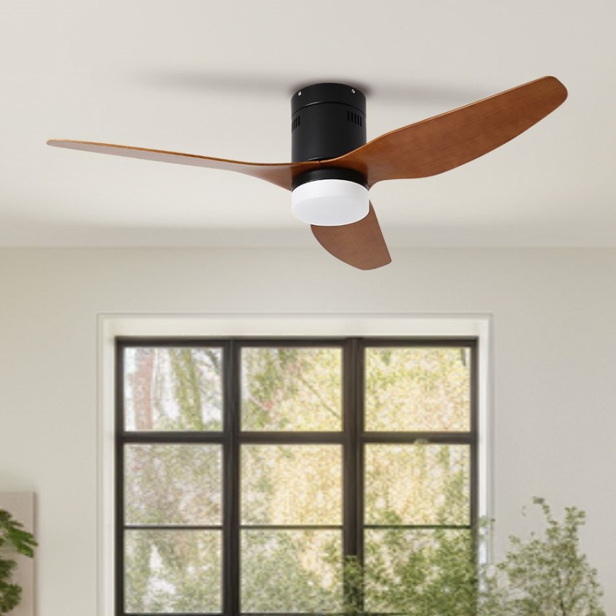 Trojlistý stropný ventilátor s drevenými lopatkami a čiernym motorom sa otáča. Je osvetlený zabudovaným bielym svetlom. Nachádza sa v miestnosti s oknom.