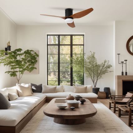 Stropný ventilátor s drevenými lopatkami a čiernym telom sa otáča v svetlej obývacej izbe s bielym gaučom, dreveným konferenčným stolíkom a dvoma izbovými rastlinami.