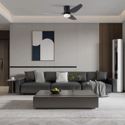 Stropný ventilátor s troma lopatkami sa otáča v moderne zariadenej obývacej izbe. Je umiestnený nad tmavosivou rozkladacou sedačkou a nízkym konferenčným stolíkom. V pozadí je stena s obrazom a drevom.