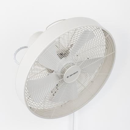 Biely nástenný ventilátor s troma lopatkami rotuje. Je pripevnený na stene, viditeľný je biely kábel. Na ventilátore je logo „ZEVENTI“.
