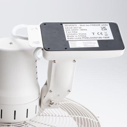 Biely nástenný ventilátor je pripevnený k stene. Jeho motor je umiestený na pohyblivom rameni. Štítok uvádza: ZEVENTI - Wall fan FREEZE white, SKU#:98730, 220-240V-50Hz, Fan: 55W, Climatic Class, Made in China, Date code: POBLE000186-1825.