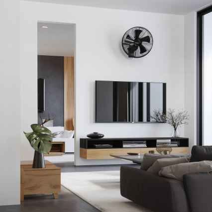 Stenový ventilátor je umiestnený nad televízorom v moderne zariadenej obývacej izbe. Televízor je umiestnený na nástennom televíznom stolíku s doplnkovými predmetmi. V pozadí je viditeľná časť spálne.