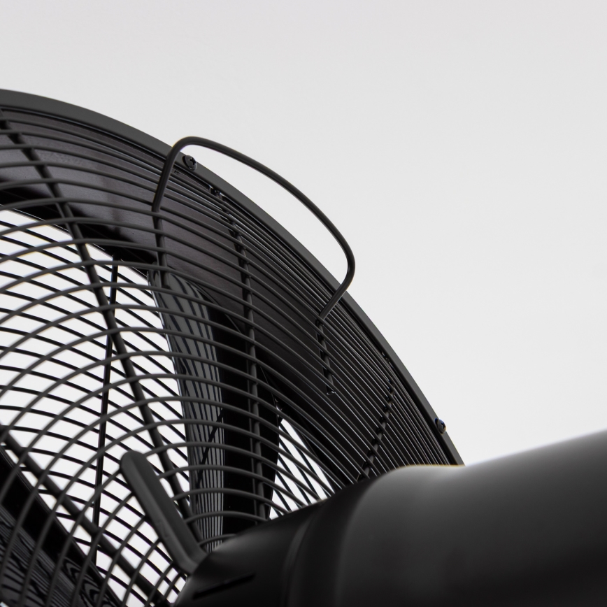 Veľký čierny ventilátor s kovovou ochrannou mriežkou a rúrkocou rukoväťou je zobrazený zblízka pred svetlým pozadím. Ventilátor je statický.