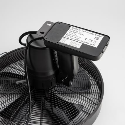 Čierny nástenný ventilátor s kovovými vrtuľami je pripojený k elektrickej sieti. Na zadnej strane je viditeľný štítok s údajmi: ZEVENTI - Wall fan FREEZE ORB, SKU#:98747, 220-240V-50Hz, Fan:55w, Climatic Class, Made in China, Date code: POBLE000189-1825. Ventilátor je umiestnený na bielom pozadí.