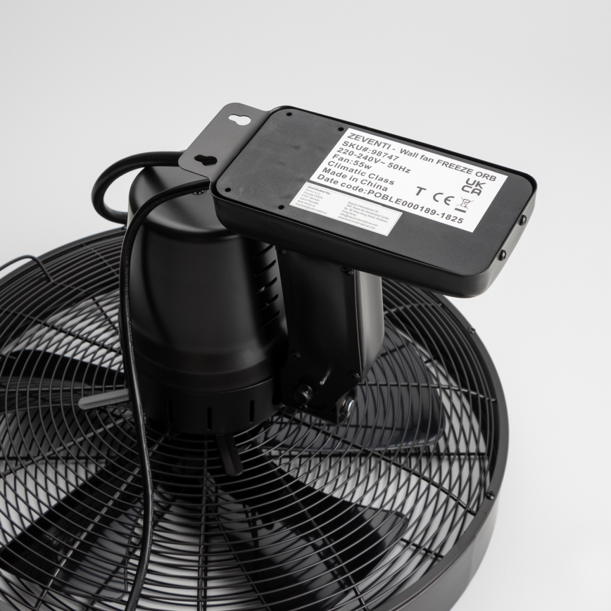 Čierny nástenný ventilátor s kovovými vrtuľami je pripojený k elektrickej sieti. Na zadnej strane je viditeľný štítok s údajmi: ZEVENTI - Wall fan FREEZE ORB, SKU#:98747, 220-240V-50Hz, Fan:55w, Climatic Class, Made in China, Date code: POBLE000189-1825. Ventilátor je umiestnený na bielom pozadí.