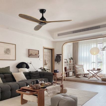 Drevený stropný ventilátor s troma lopatkami rotuje v obývacej izbe zariadenej v minimalistickom štýle s pohovkou, konferenčným stolíkom a doplnkami. Izba má svetlé steny a veľké okná.
