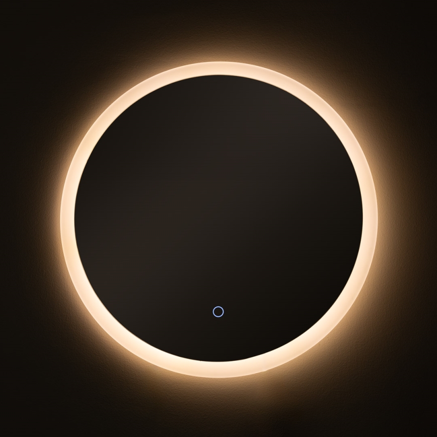 Okrúhle zrkadlo je osvetlené LED diódami po obvode. Zrkadlo je umiestnené na tmavej stene. V strede zrkadla je malé, svetlomodré tlačidlo.